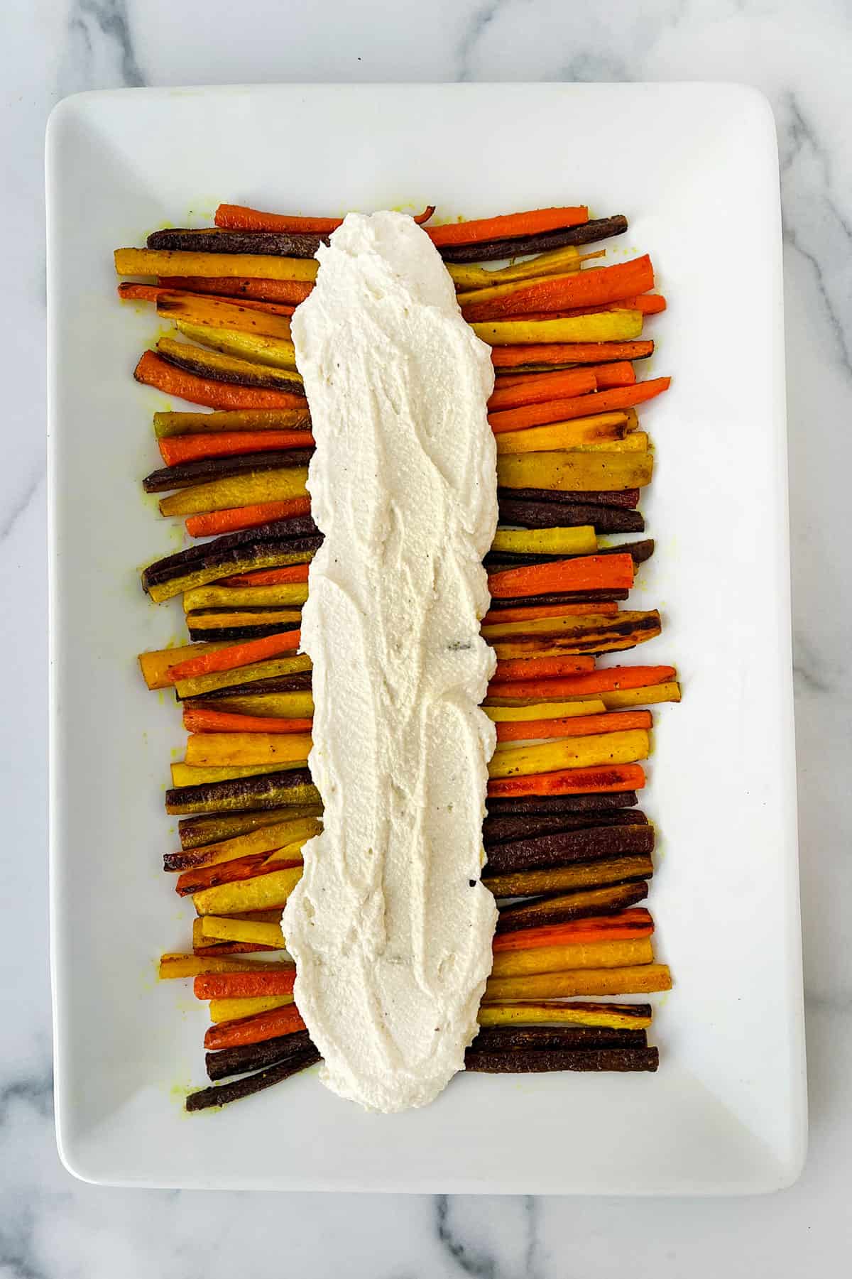 Roasted rainbow carrots sticks arranged in two rows on a rectangular platter with whipped feta spread down the middle between the rows.