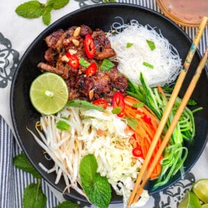 Vietnamese noodle salad bowl - bun this nuong - with caramelized pork, vermicelli rice noodles, shredded carrots, cabbage and cucumbers, bean sprouts and half a lime, chopsticks resting on the bowl.