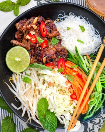 Vietnamese noodle salad bowl - bun this nuong - with caramelized pork, vermicelli rice noodles, shredded carrots, cabbage and cucumbers, bean sprouts and half a lime, chopsticks resting on the bowl.