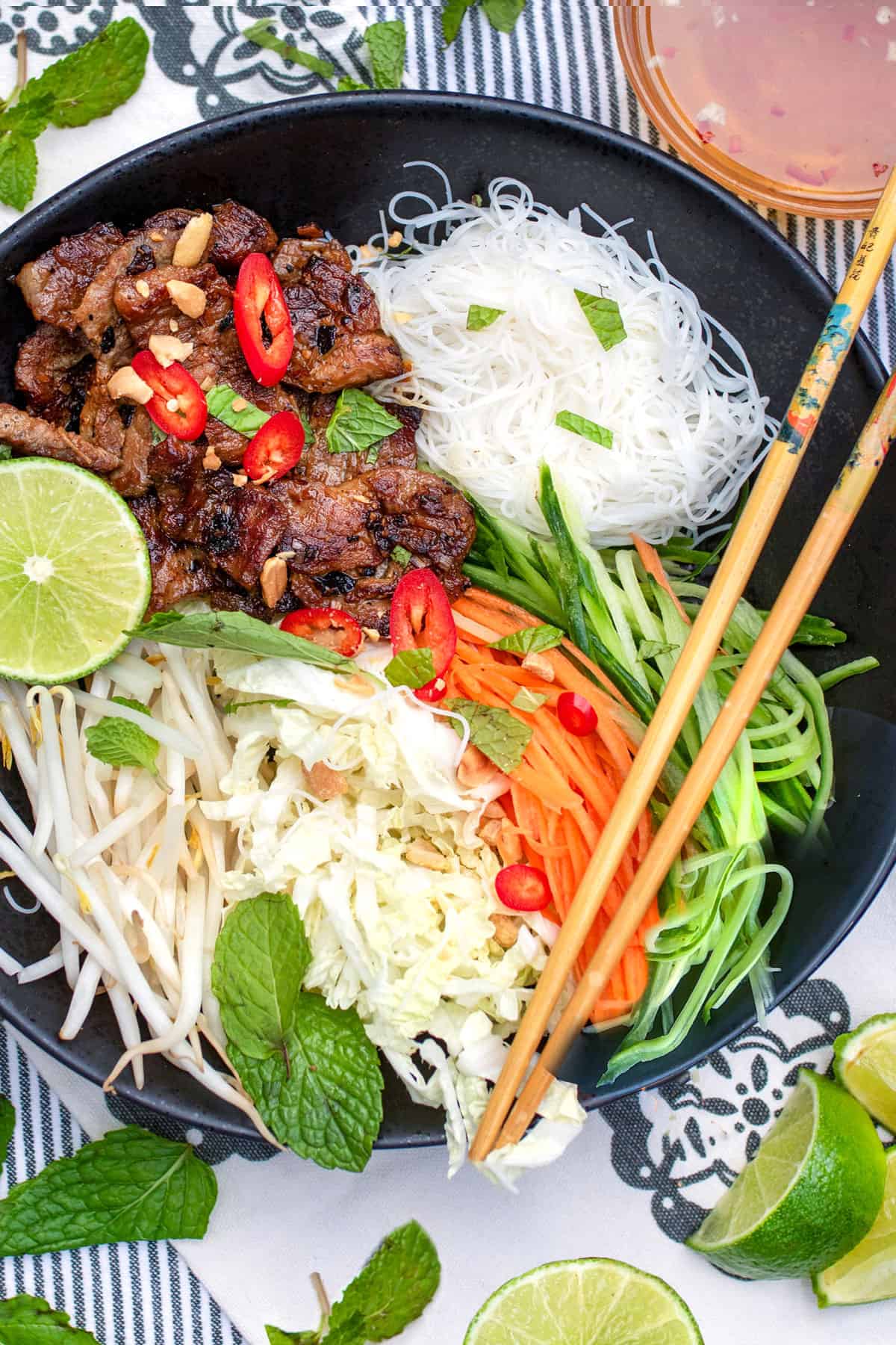 Bowl filled with piles of ingredients: vermicelli rice noodles, caramelized pork, shredded carrots, cabbage and cucumbers, and bean sprouts. Chopsticks resting on the edges of the bowl.