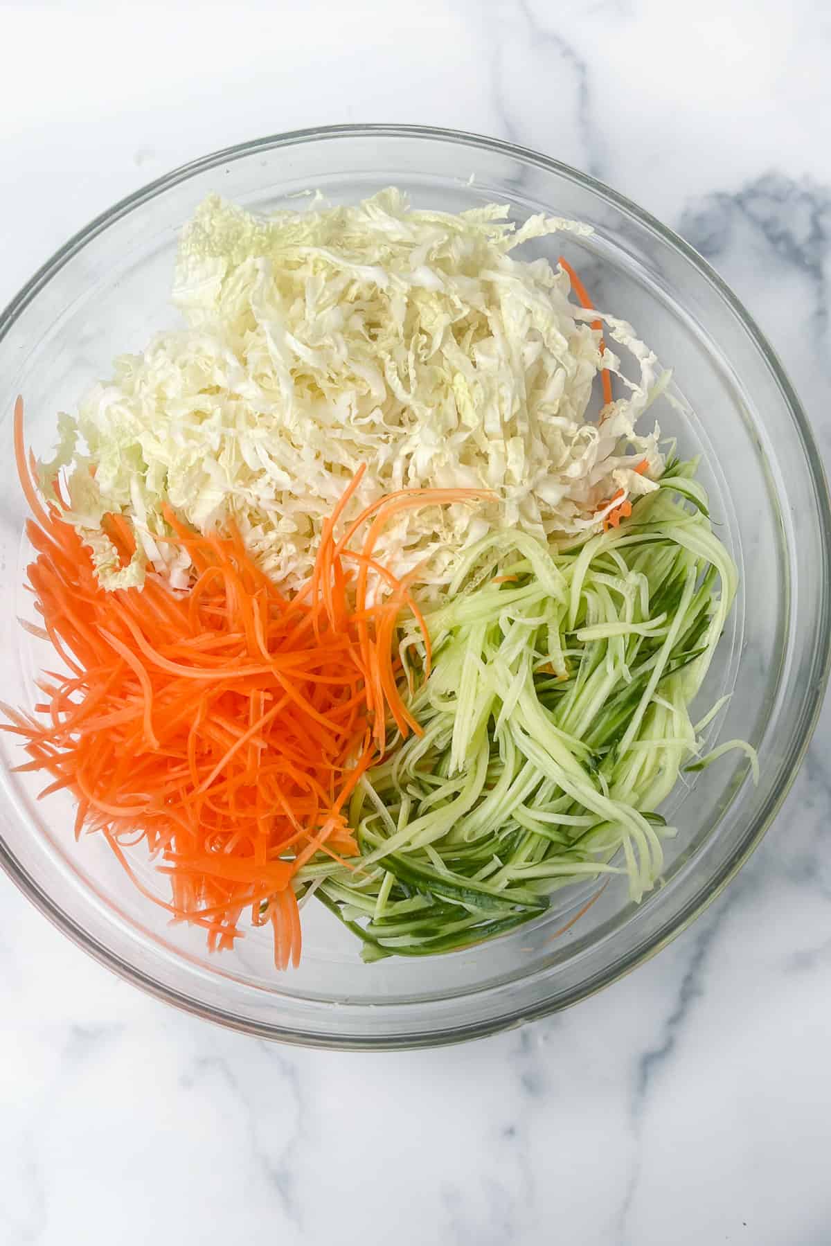 Glass bowl with a pile of shredded carrots, pile of shredded napa cabbage and pile of shredded cucumbers.