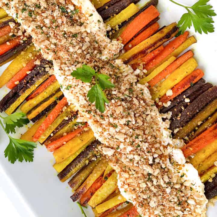 Two rows of roasted rainbow carrots arranged on a rectangular platter with whipped feta down the center and toasted bread crumbs sprinkled on top.