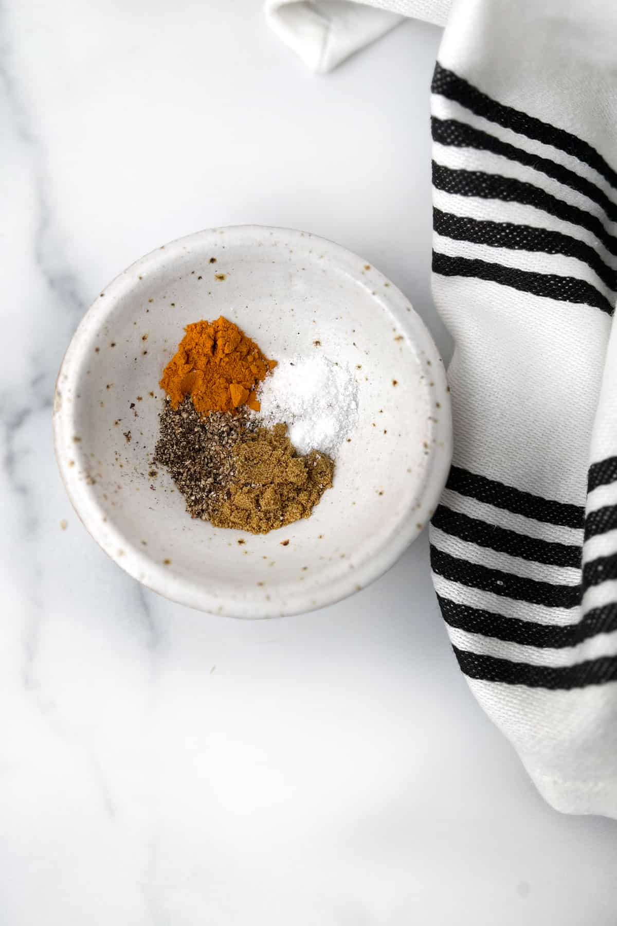 small white bowl with 4 spices in piles: turmeric, cumin, salt and pepper.