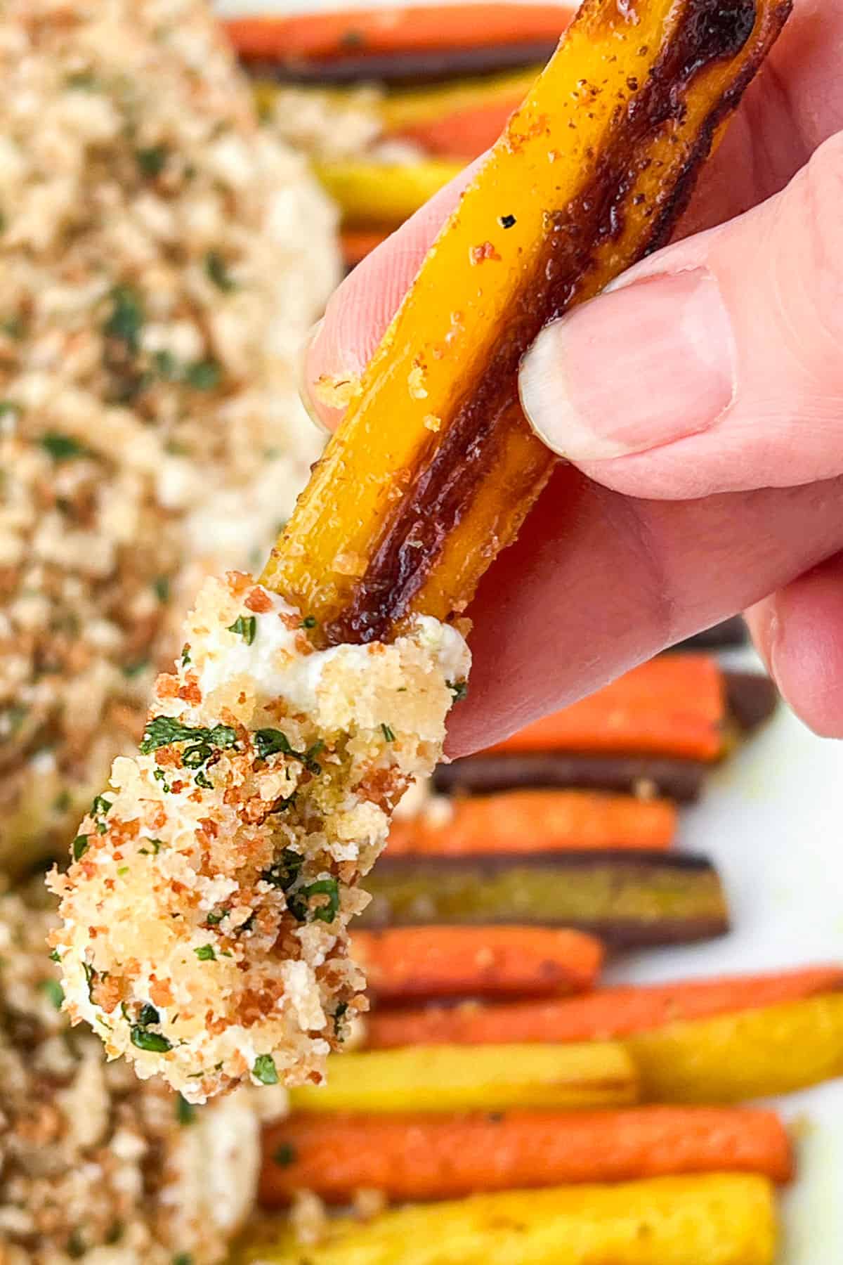 Hand holding one yellow roasted carrot with the tip covered in creamy whipped feta and toasted breadcrumbs.