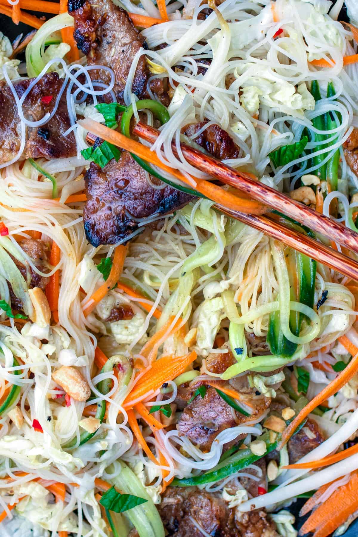 A tangle of thin rice noodles, shredded carrots, cucumber and cabbage, and caramelized pork, with chopsticks holding one bite-sized piece of pork.