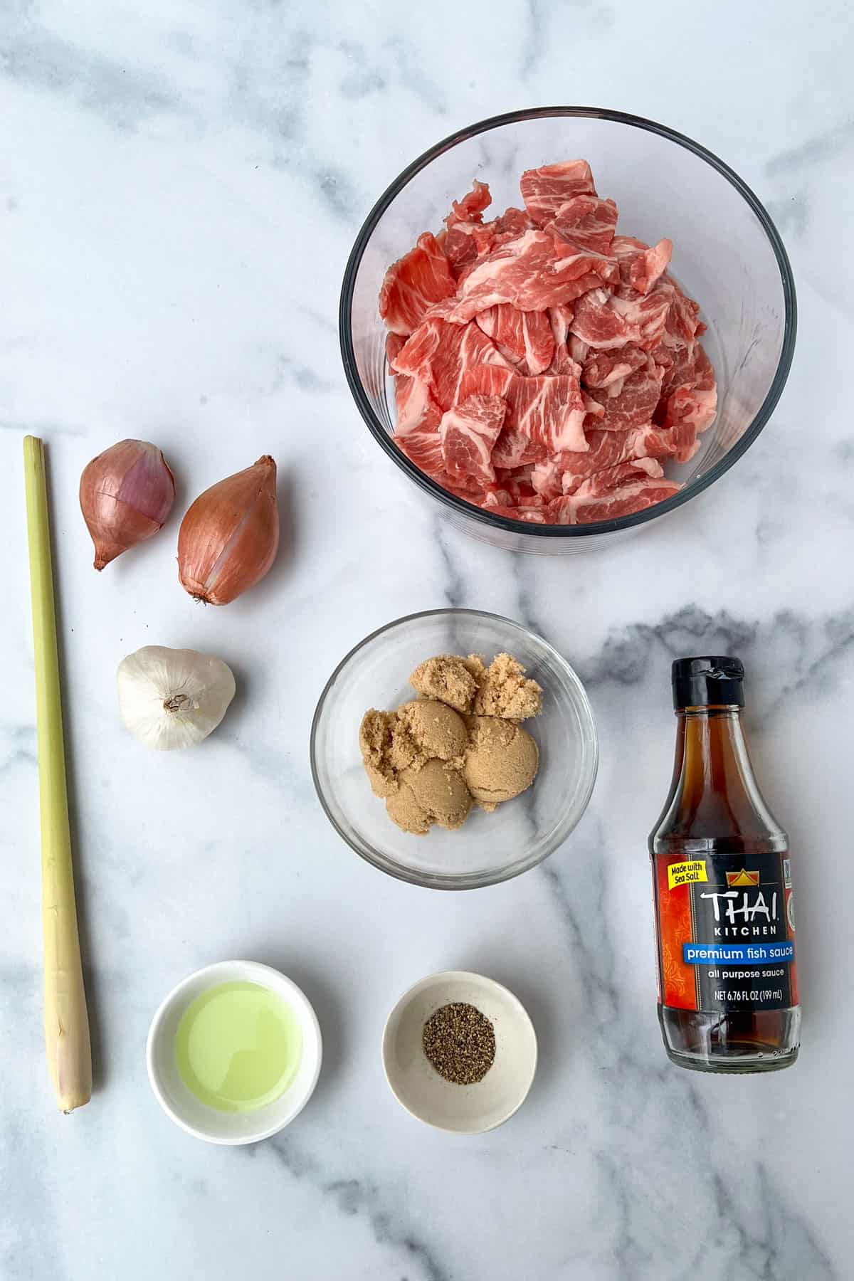 Ingredients on a marble countertop: bowl of thinly sliced pork shoulder, two shallots, a lemongrass stalk, a small bowl of cracked pepper, small bowl of oil, bowl of light brown sugar, bottle of fish sauce