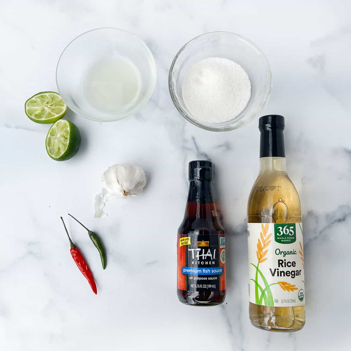 Ingredients on a marble counter: two halves of a squeezed lime, two small Thai chilis, bulb of garlic, bowl of white sugar, bowl of lime juice, bottle of fish sauce and bottle of rice vinegar.