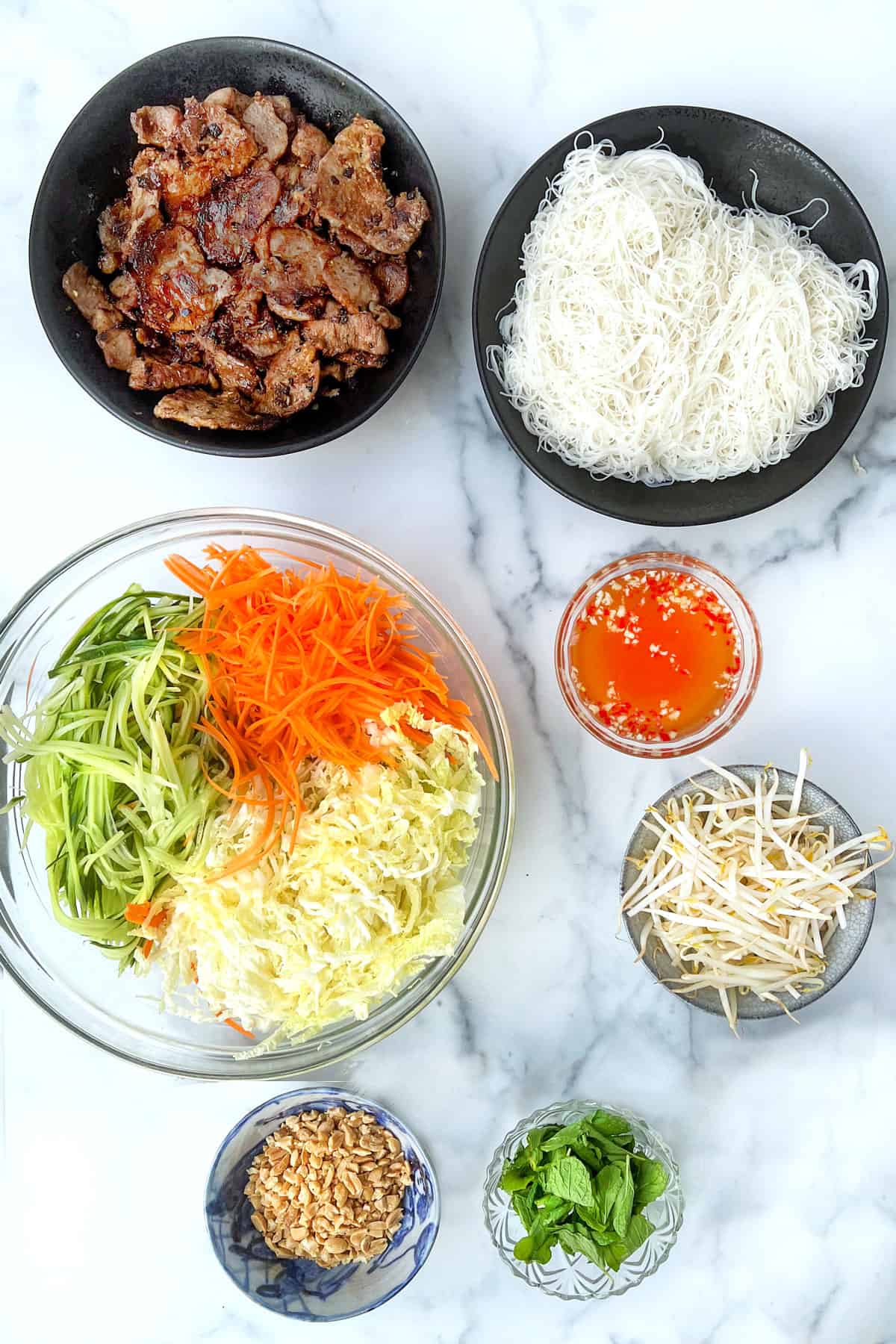 bowls of ingredients on a marble counter: caramelized pork slices, thin vermicelli rice noodles, shredded carrots, cucumbers and cabbage in a glass bowl, bean sprouts, chopped peanuts, mint leaves, and vietnamese chili garlic dipping sauce.