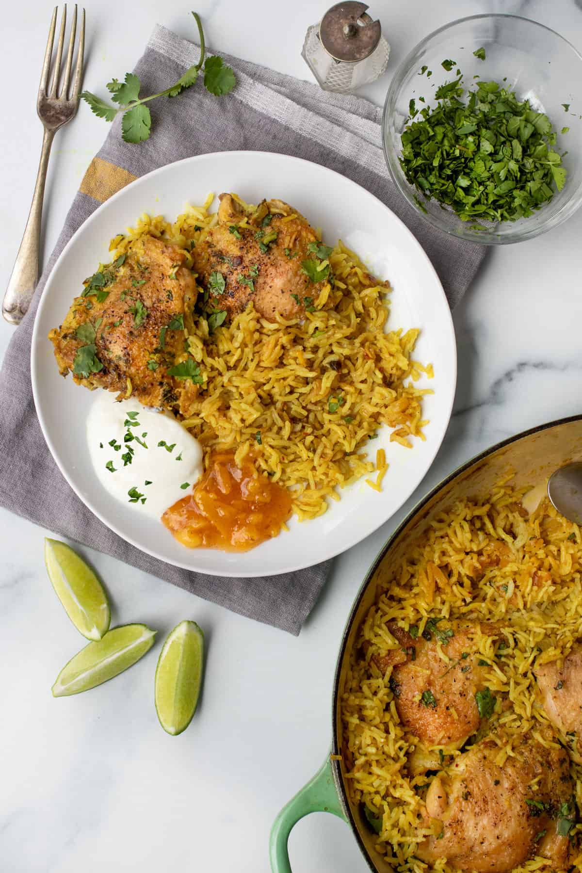 A dinner plate with two baked chicken thighs on a bed of turmeric colored rice with a small blob of yogurt and a small blob of mango chutney. The pot of chicken and rice to the side and some lime wedges and a fork on the side.