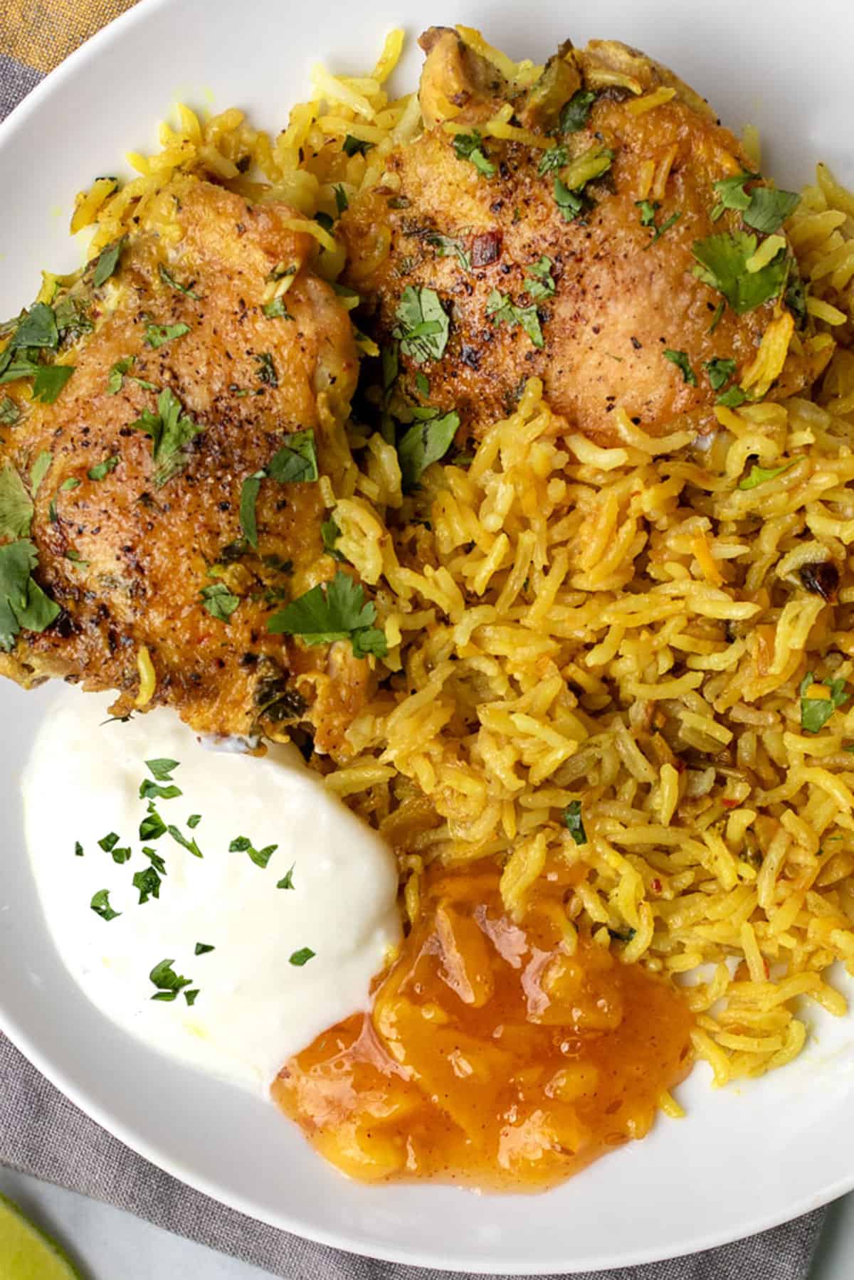 Two golden brown baked chicken thighs next to a bed of fluffy turmeric colored rice, all topped with chopped cilantro, with a small blob of yogurt and small blob of mango chutney on the plate.