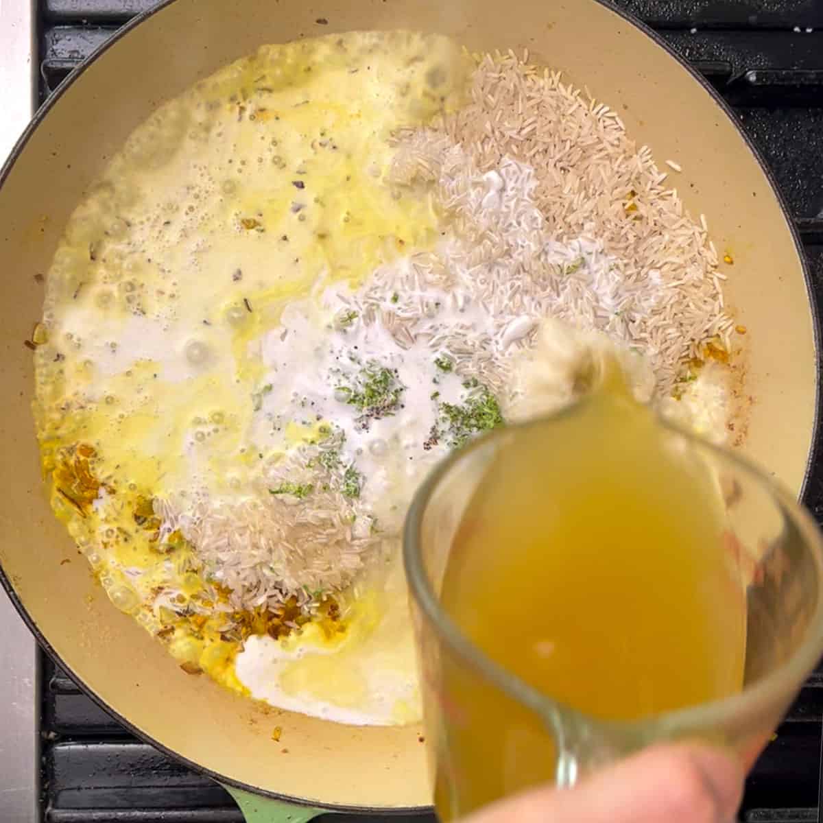Shallow dutch oven with aromatics and basmati rice and lime zest and coconut milk and chicken stock being poured in from a glass measuring cup.