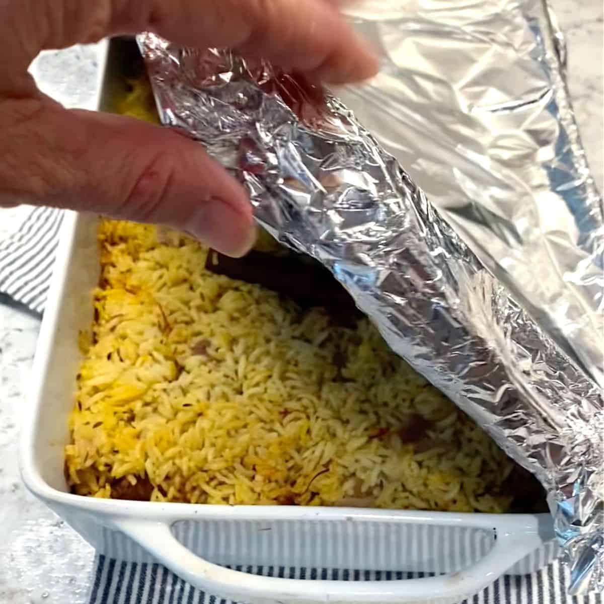 Casserole pan filled with lamb biryani, topped with foil with a corner of the foil lifed so the rice layer is seen beneath.