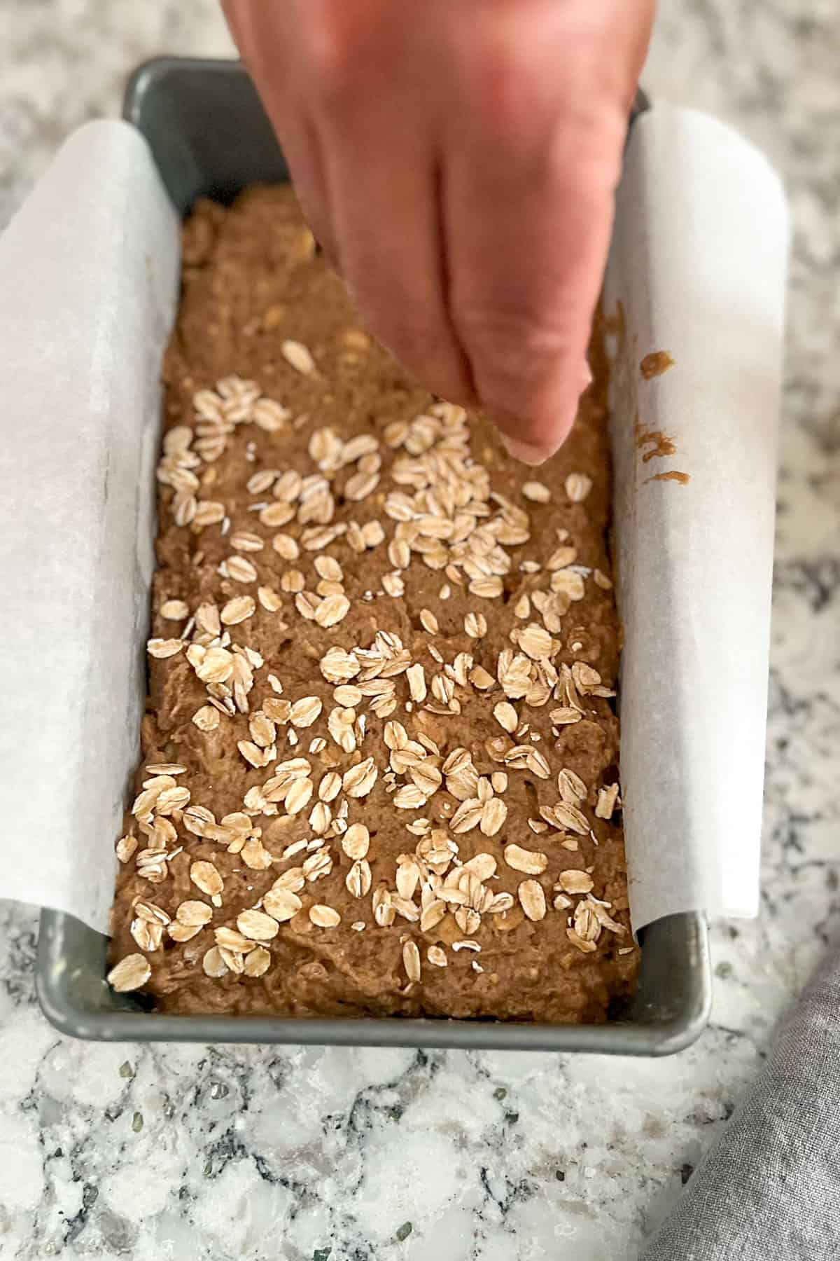 Hand sprinkling rolled oats over bread dough in loaf pan.