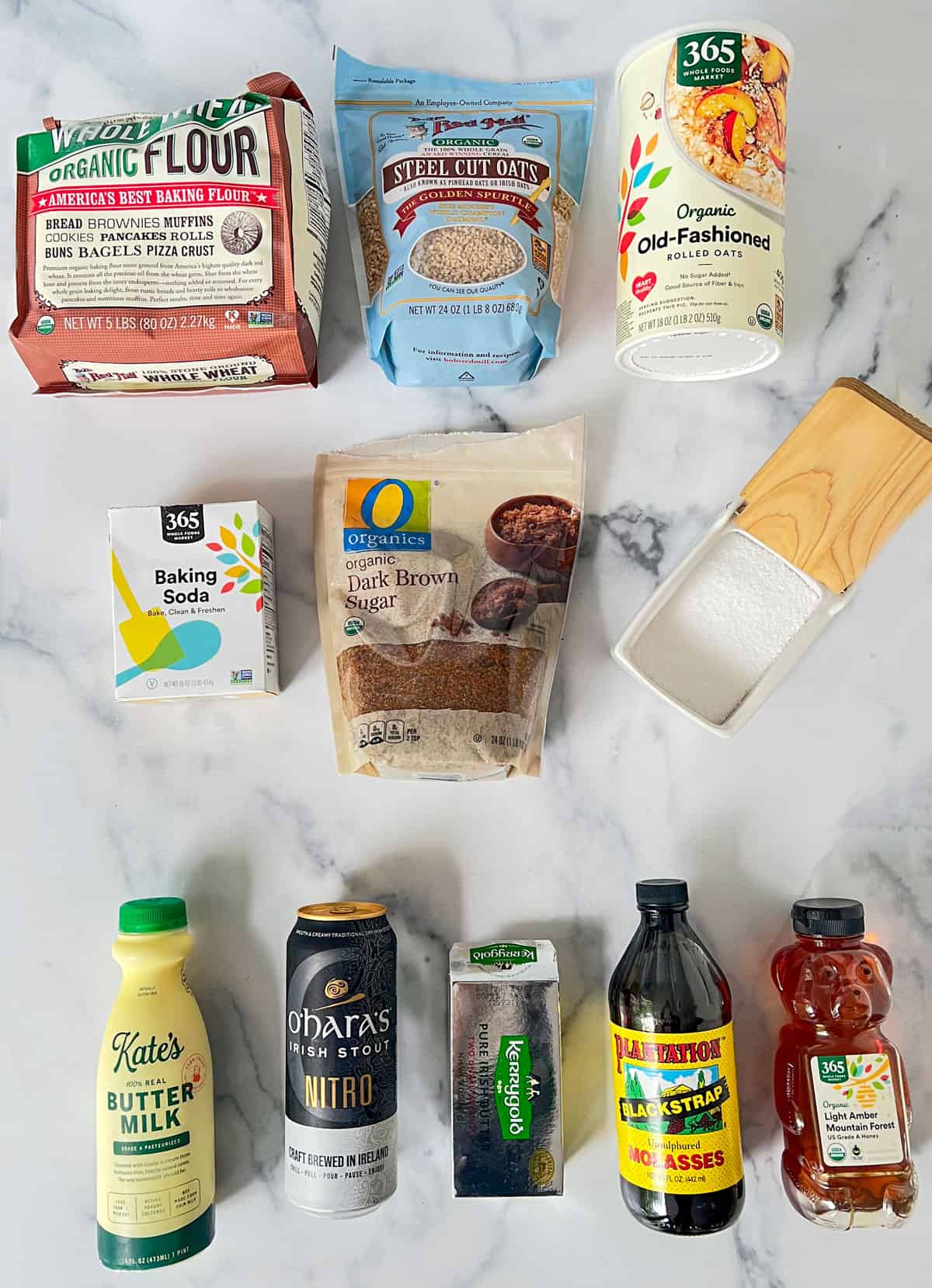 Ingredients in their containers laid out on a marble countertop: flour, two types of oats, baking soda, brown sugar, salt, buttermilk, ale, Irish butter, molasses, honehy