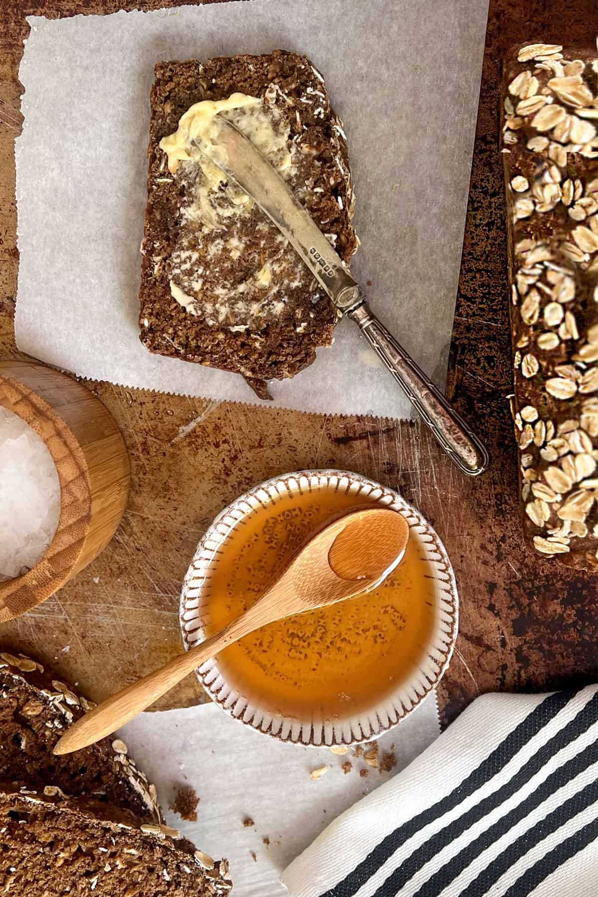 a slice of brown bread being buttered next to a small bowl of honey with a wooden spoon and a wooden container of sea salt.