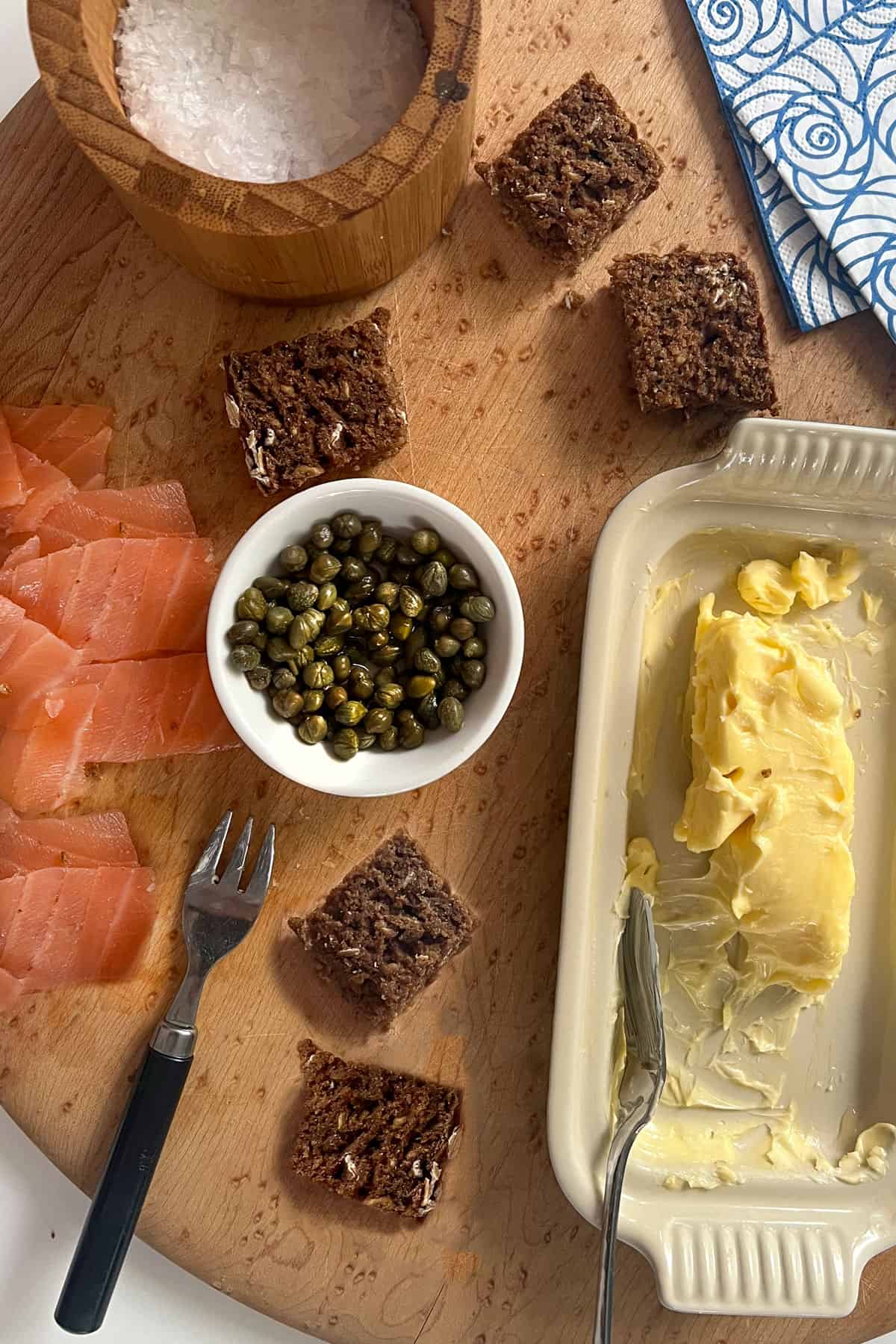 Wooden platter topped with small squares of brown bread, small slices of smoked salmon, a tiny bowl of capers, a container of sea salt and a butter dish with softened buttter.