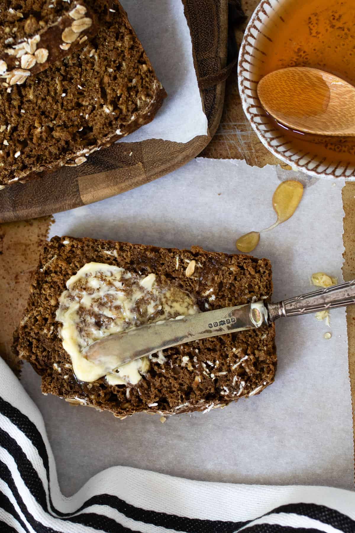 brown bread being slathered with butter by a small butterknife, a bowl of honey to the side.