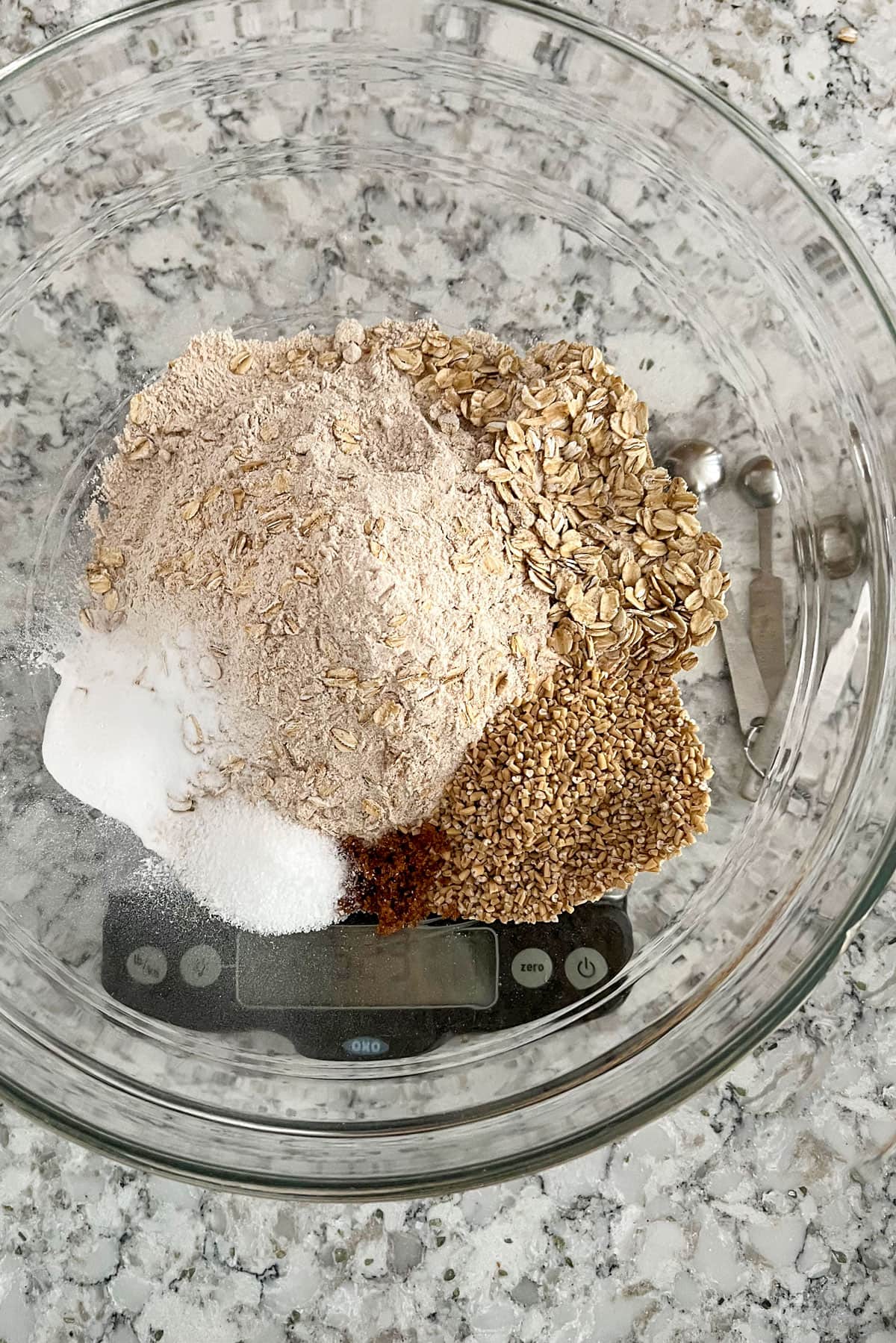 Glass mixing bowl on a kitchen scale, filled with rolled oats, steel cut oats, whole wheat flour, brown sugar, salt and baking soda.