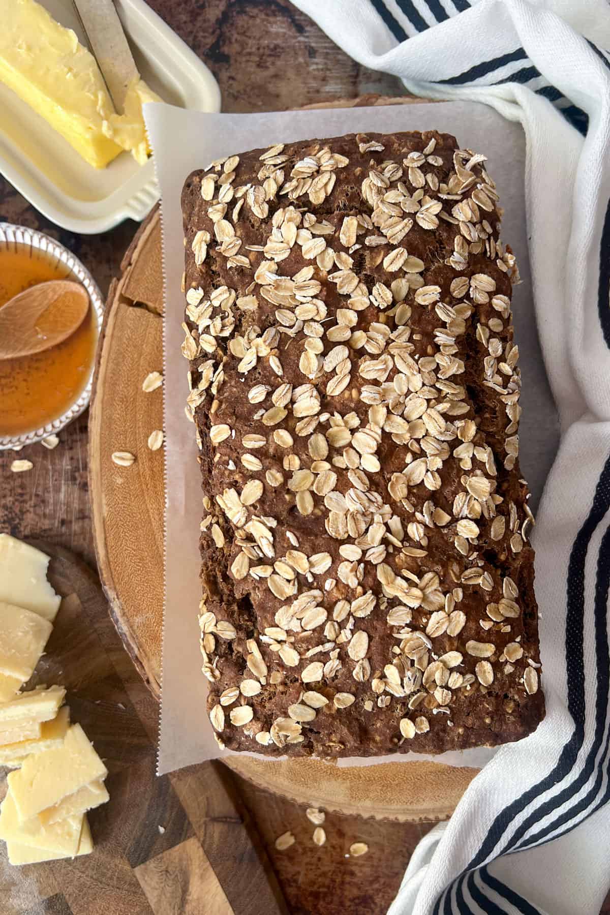 Loaf of brown bread topped with rolled oats on a wooden platter surrounded by butter, honey and sliced cheddar cheese