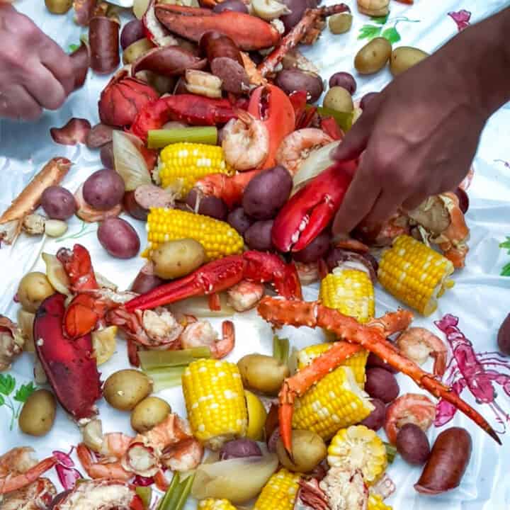 Seafood boil with lobsters, shrimp, clams, crab legs, corn, and potatoes, poured out onto a lobster-print plastic tablecloth - hands grabbing seafood.