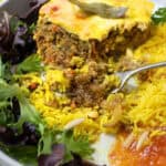 Plate topped with a slice of bobotie, meat casserole with custard on top, yellow rice and salad.