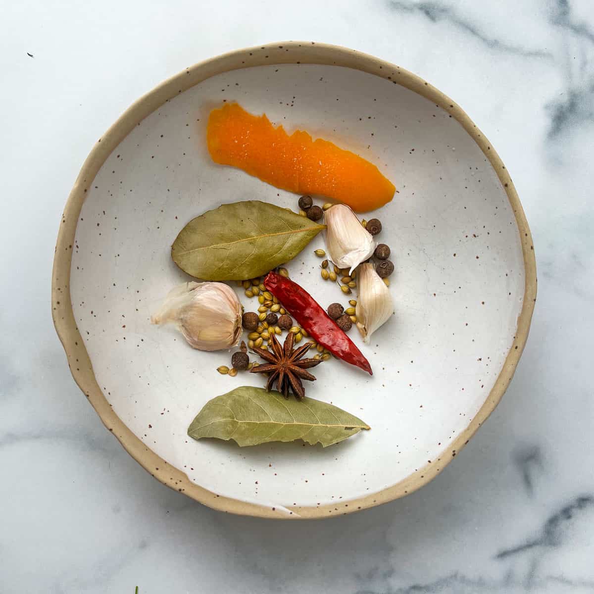 assorted whole spices in a small bowl with a piece of orange peel and 3 unpeeled garlic cloves and a dried red chili.