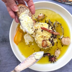 round soft cheese in shallow bowl surrounded with olive oil, herbs and whole spices. Hand holding a baguette slice that has been spread with some of the cheese.