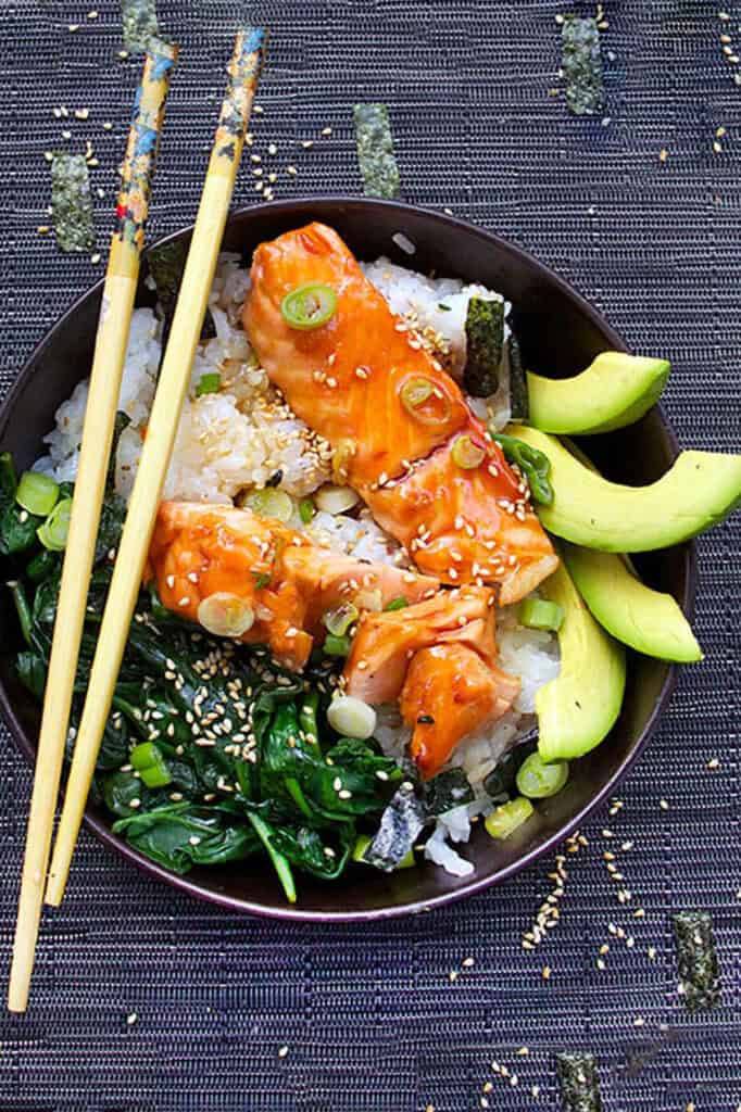 bowl with two pieces of broiled salmon on a bed of rice with sautéd spinach, slices of avocado and sesame and nori confetti sprinkled around.
