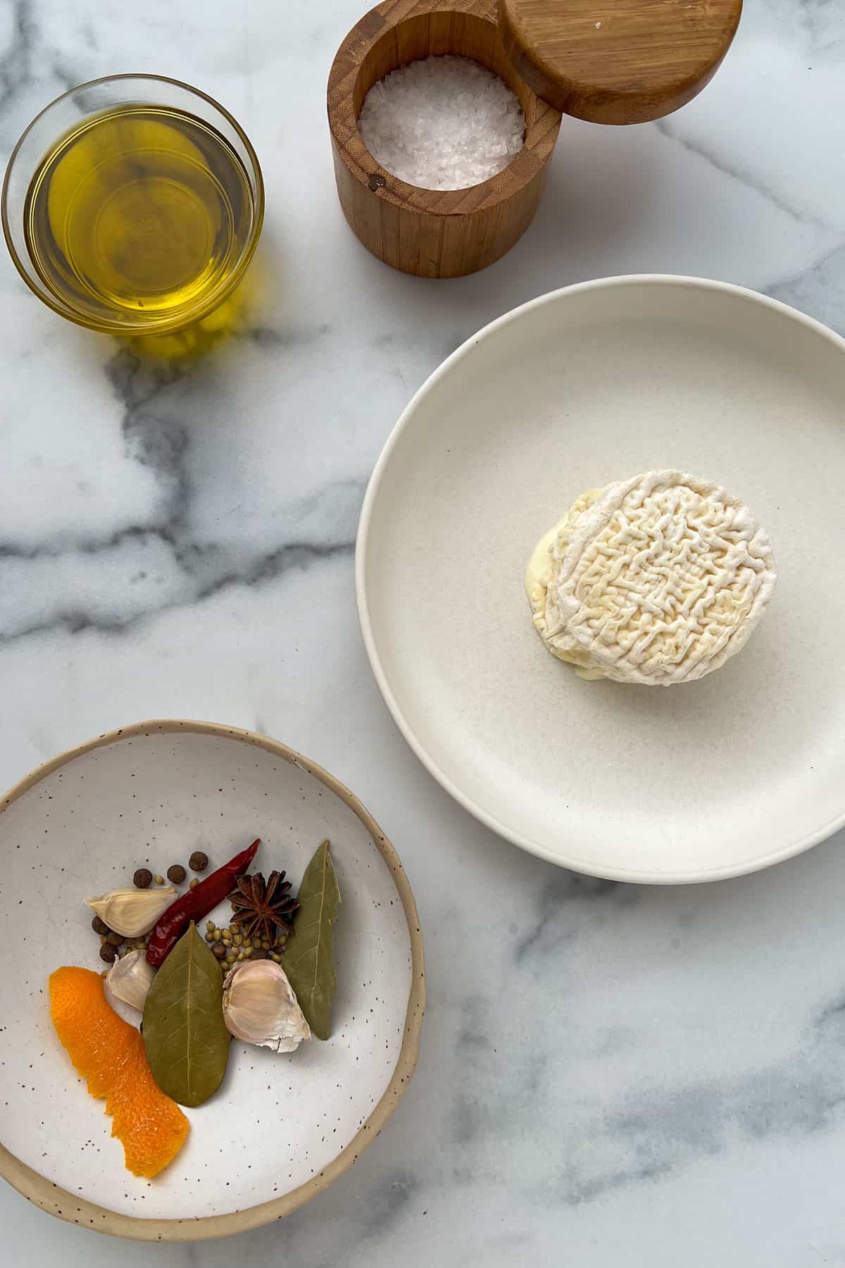 Marble counter with bowl of whole spices, small har of olive oil, salt cellar, and rimmed plate with a wheel of La Tur cheese in the center.