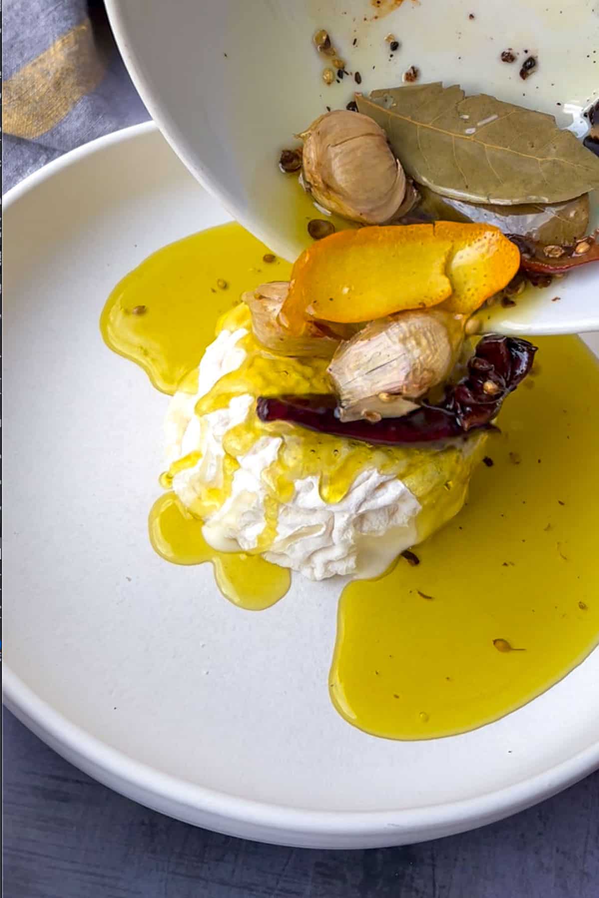 Herb and garlic infused olive oil being poured from a small porcelain pot over a wheel of cheese, with all the whole spices falling down onto the cheese and onto the serving plate.