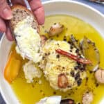 round soft cheese in shallow bowl surrounded with olive oil, herbs and whole spices. Hand holding a baguette slice that has been spread with some of the cheese.