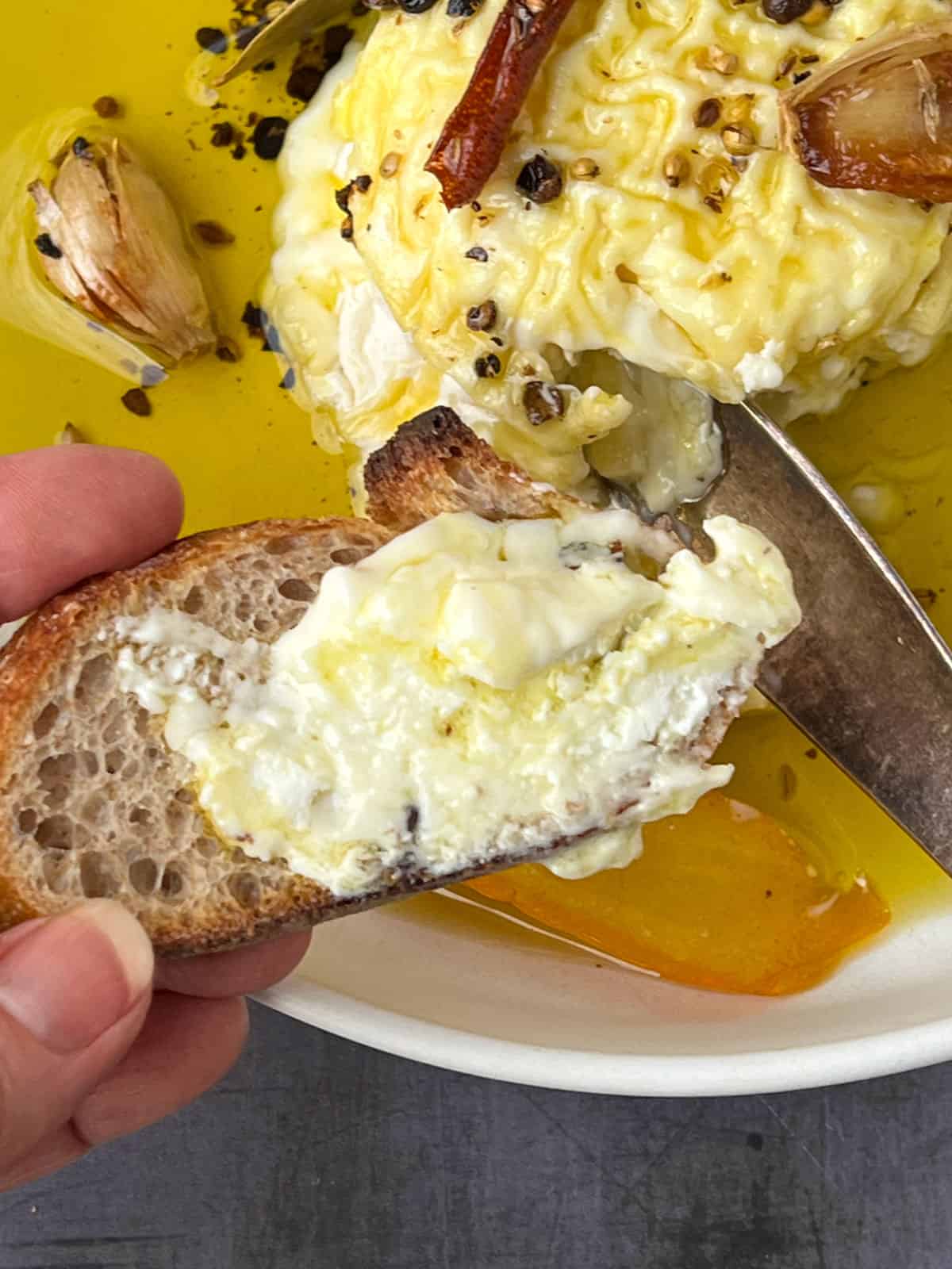 Hand holding a piece of crostini slathered with creamy cheese and olive oil, the wheel of cheese and cheese knife in the background.