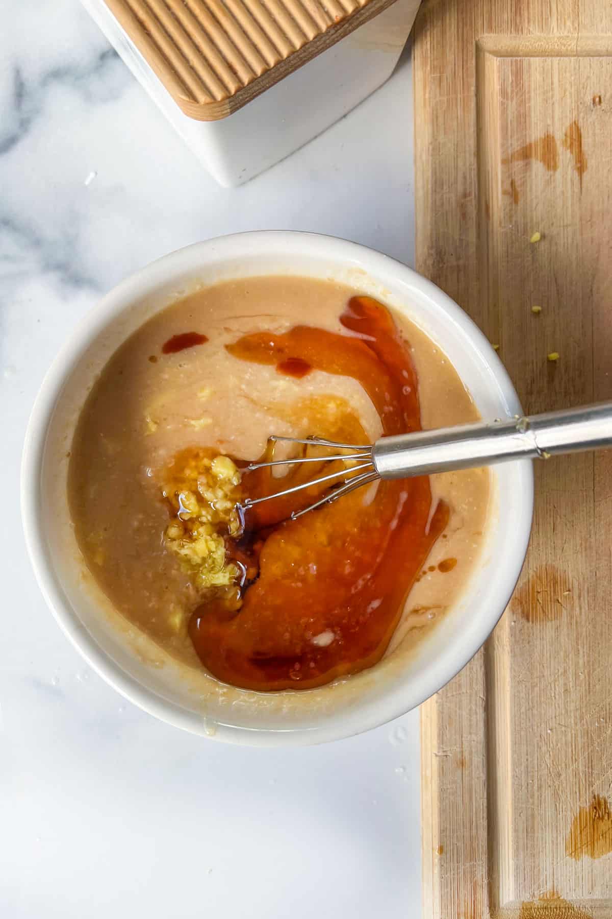 White bowl filled with dressing ingredients: creamy miso with sesame oil on top and chopped ginger, all being stirred together with a small whisk.