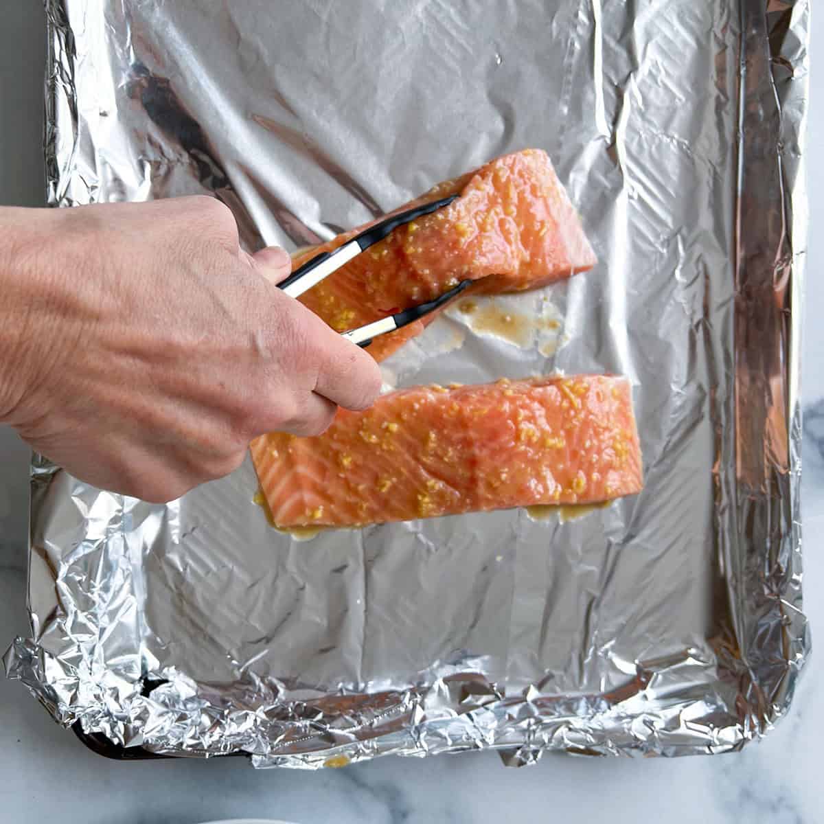 Two raw salmon fillets topped with miso ginger dressing on a foil lined baking sheet, one piece of salmon being lowered onto the baking sheet with tongs.