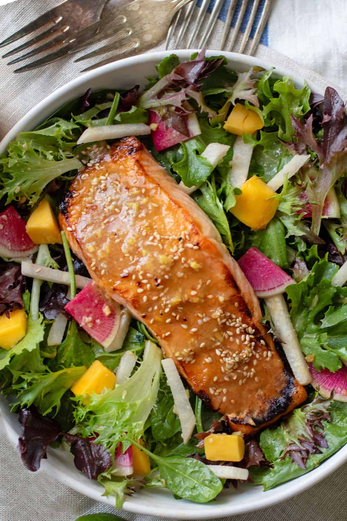 A broiled miso salmon fillet topped with miso dressing and sesame seeds on a bed of salad greens with jicama matchsticks, mango chunks and sliced watermelon radishes.