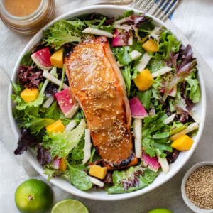 A broiled miso salmon fillet topped with miso dressing and sesame seeds on a bed of salad greens with jicama matchsticks, mango chunks and sliced watermelon radishes.