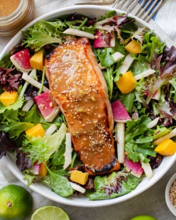 A broiled miso salmon fillet topped with miso dressing and sesame seeds on a bed of salad greens with jicama matchsticks, mango chunks and sliced watermelon radishes.