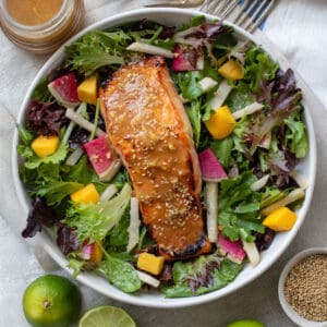 A broiled miso salmon fillet topped with miso dressing and sesame seeds on a bed of salad greens with jicama matchsticks, mango chunks and sliced watermelon radishes.