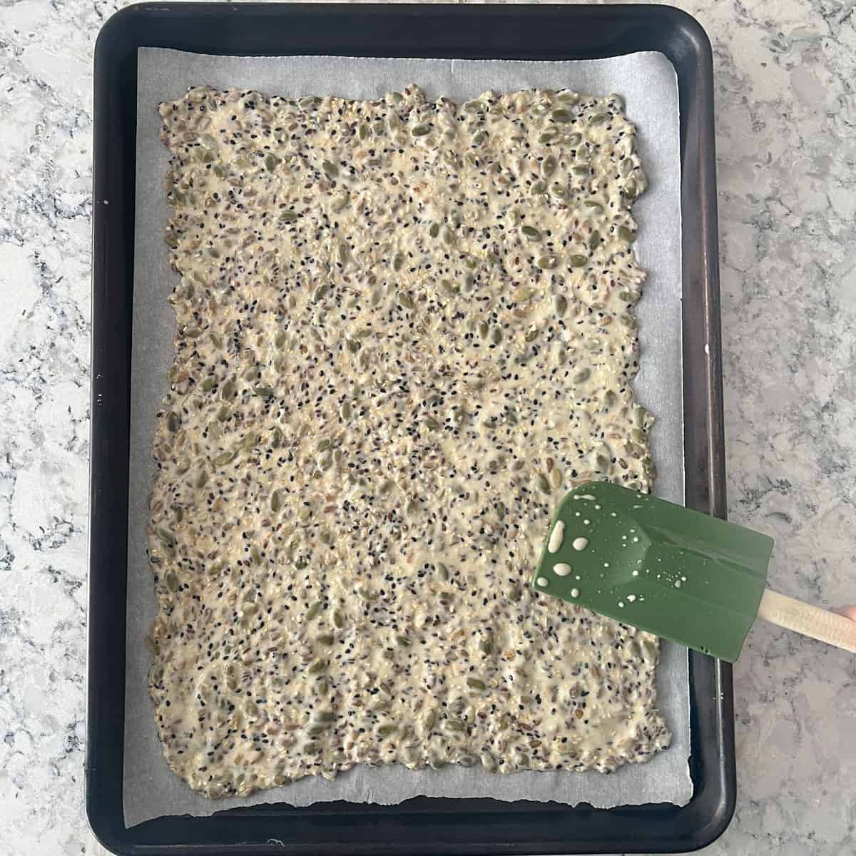 Seed cracker dough spread out into a thin rectangle on a parchment lined rimmed baking sheet with a green silicone spatula being used to finish spreading.