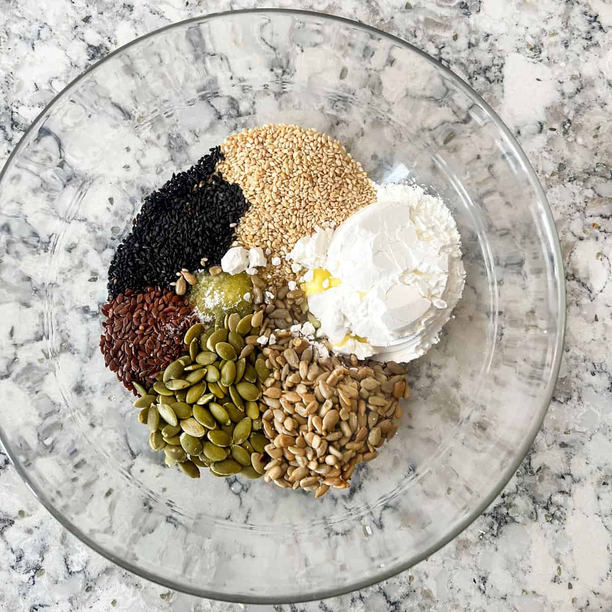 Glass mixing bowl with piles of ingredients: sesame seeds, pumpkin seeds, flax, seeds, sunflower seeds, cornstarch, salt, olive oil poured in tthe middle.