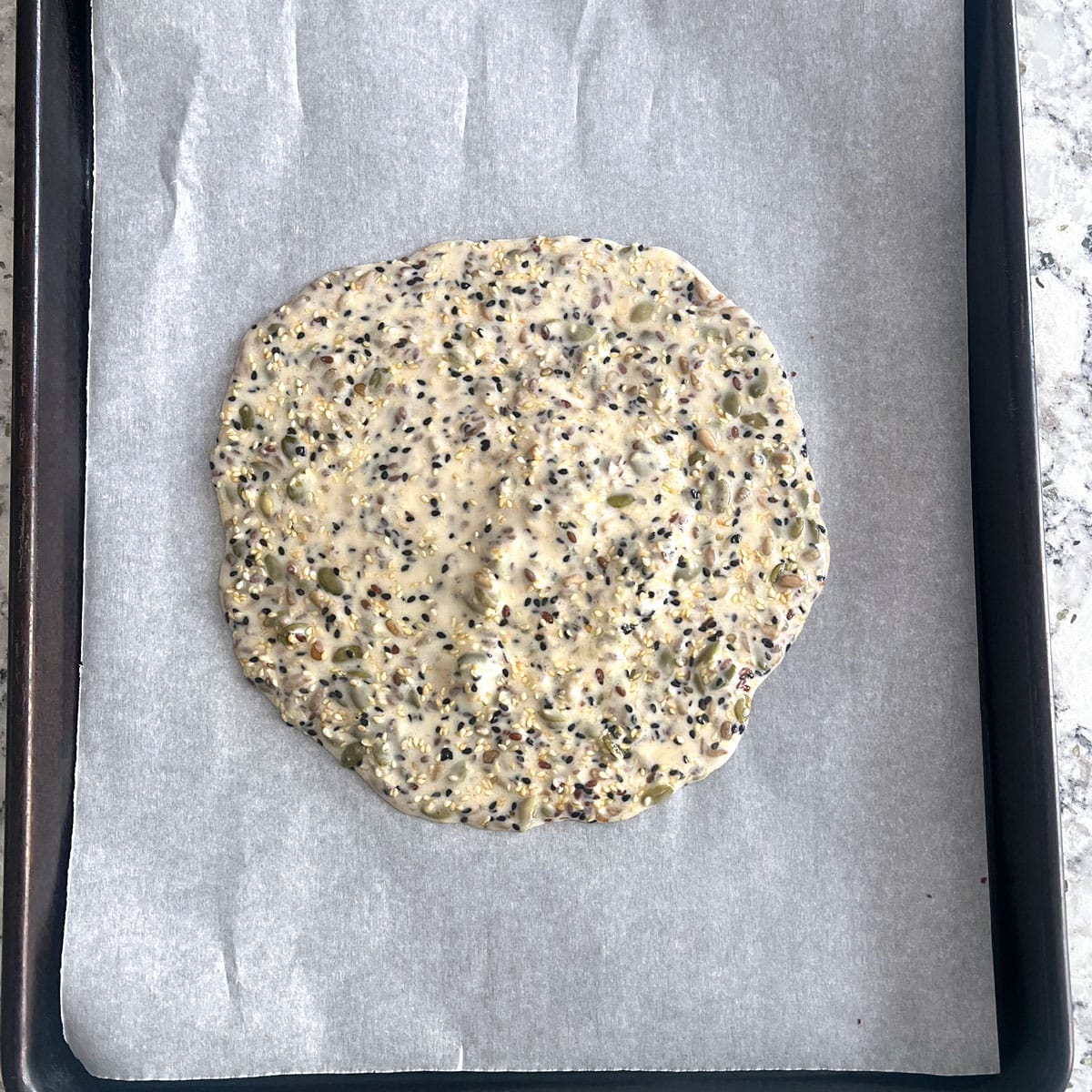 Seed cracker dough in the middle of a parchment lined rimmed baking sheet.