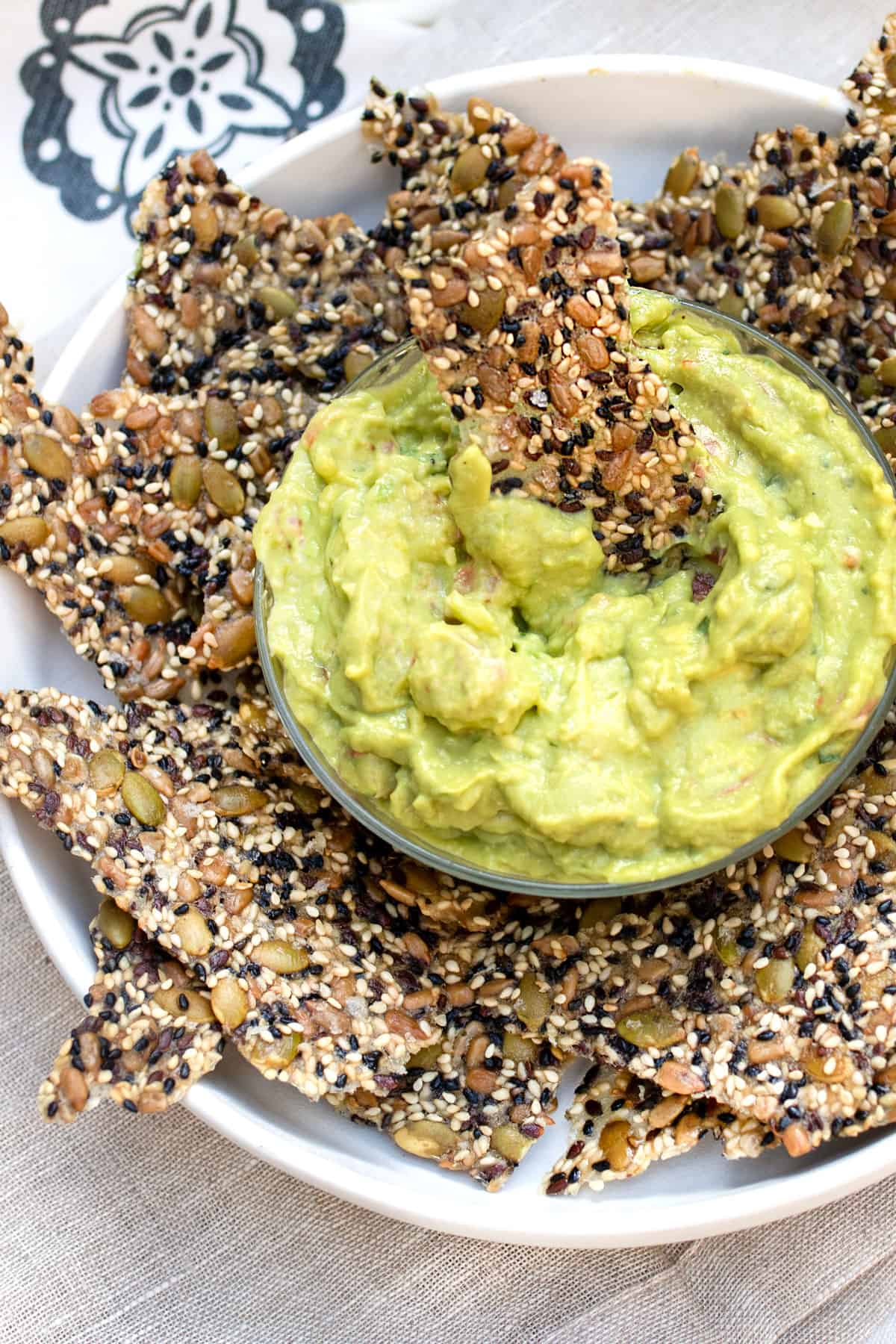 A bunch of seeded crackers arranged on a rimmed serving platter with a small bowl of guacamole in the middle and a cracker in the center of the guac.