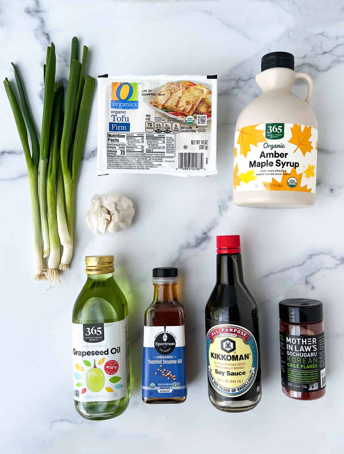 Ingredient laid out on a marble countertop: bunch of scallions, box of tofu, bottle of maple syrup, soy sauce, vegetable oil, sesame oil, container of gochugur flakes and a whole bulb of garlic.