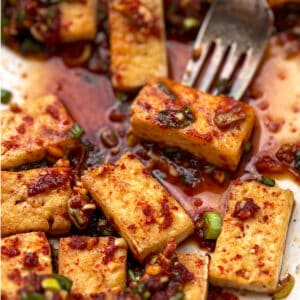bite-sized rectangular pieces of pan fried tofu in rich sauce with chopped scallions and a fork holding one piece of tofu.