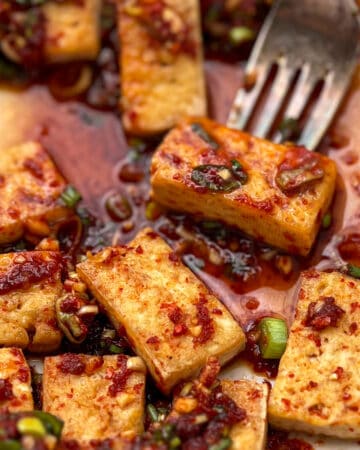 bite-sized rectangular pieces of pan fried tofu in rich sauce with chopped scallions and a fork holding one piece of tofu.