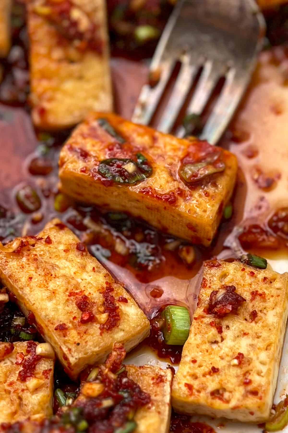 bite-sized rectangular pieces of pan fried tofu in rich sauce with chopped scallions and a fork holding one piece of tofu.
