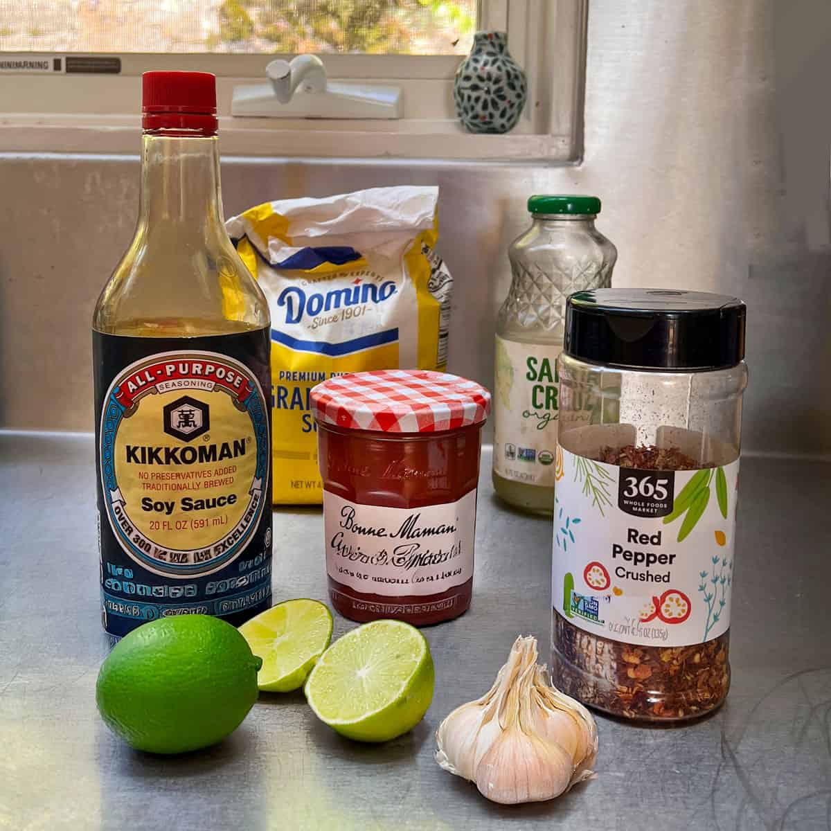 ingredients on a stainless steel countertop: bottle of soy sauce, jar of apricot preserves, 2 limes, bag of sugar, jar of crushed red pepper flakes, whole bulb of garlic.