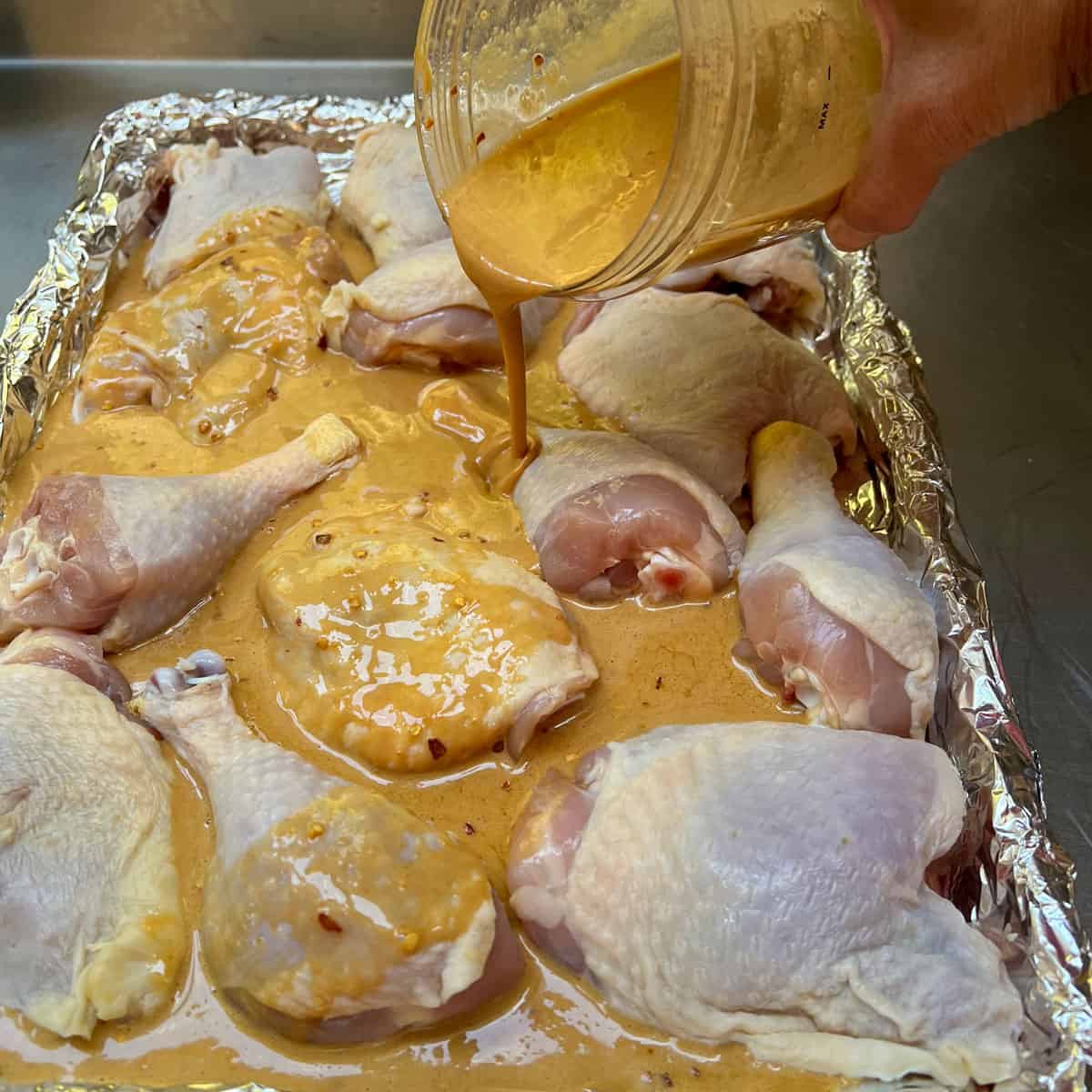 Hand holding container of apricot glaze mixture and pouring it over raw bone-in, skin-on chicken thighs and drumsticks on a foil-lined rimmed baking sheet.  