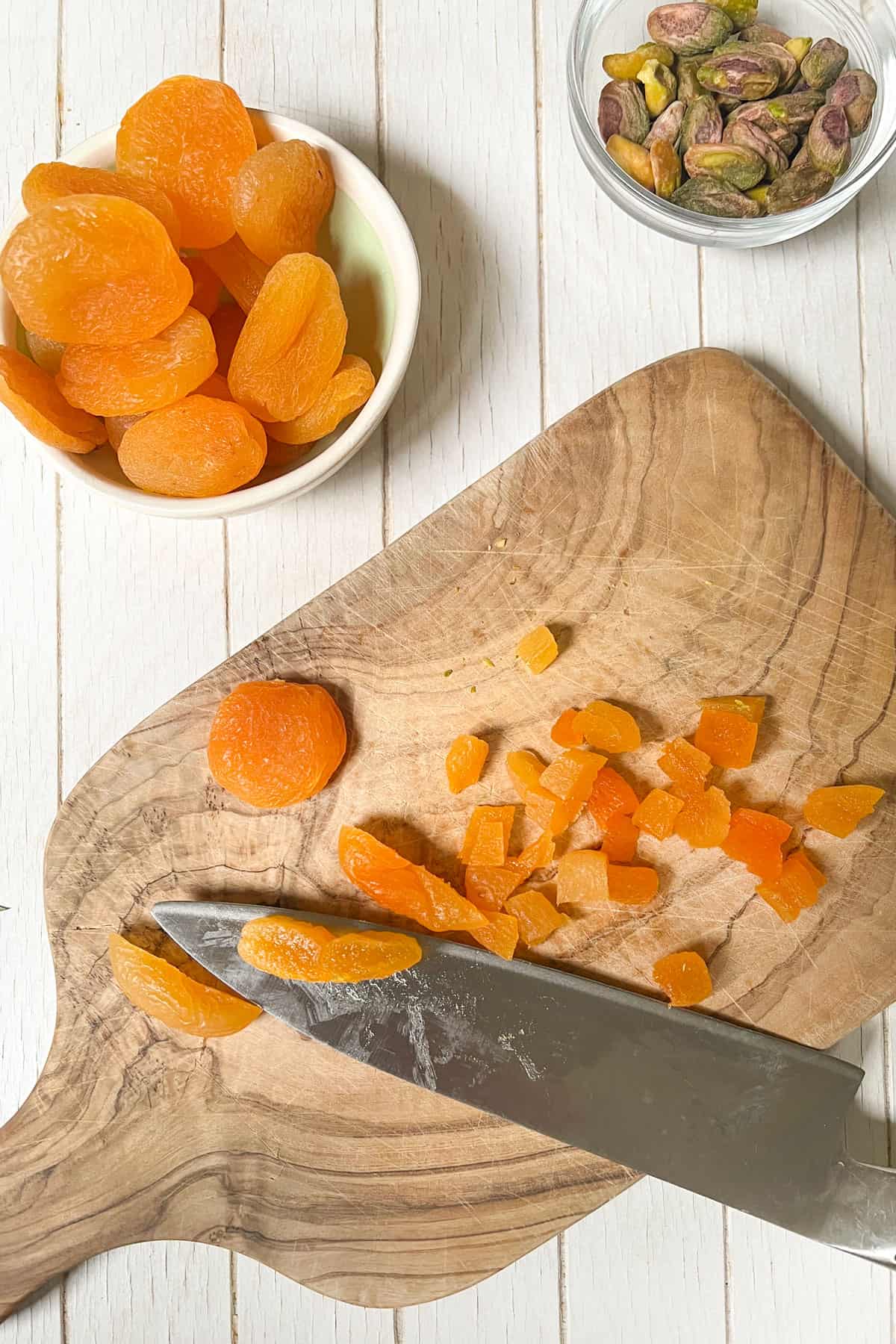 dried apricots in a small bowl and a couple getting diced on a small wooden cutting board next to a small glass bowl of toasted pistachios.
