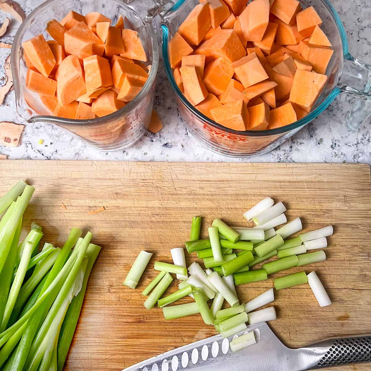 two large measuring cups filled with cubed sweet potatoes next to a cutting board with a knife and cut up scallions.